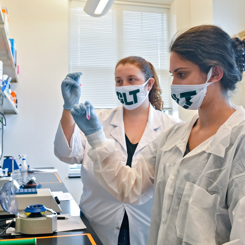 Female students wearing white lab coats with Charlotte masks in lab on campus